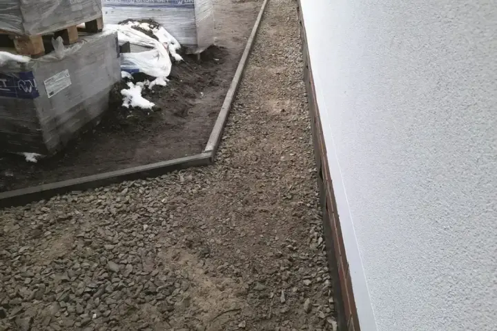Ułożona opaska przeciwwysadzinowa z kruszywa wokół budynku, obok palety z materiałami budowlanymi.