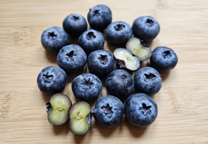 Kusząca garść borówek amerykańskich odmiany 'Bluecrop' i 'Duke' na drewnianej desce. Dwie jagody przekrojone ukazują soczysty, zielonkawy miąższ.