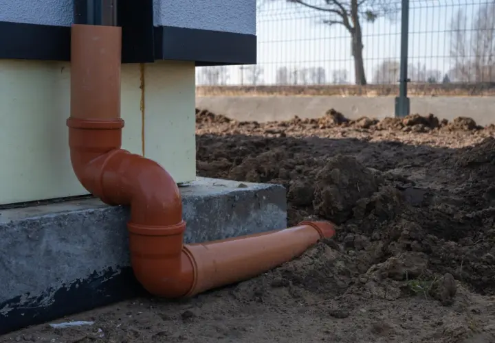 Pomarańczowe rury do odprowadzenia wody z rynien, połączone kolankami, biegną od ściany budynku w kierunku ziemi.