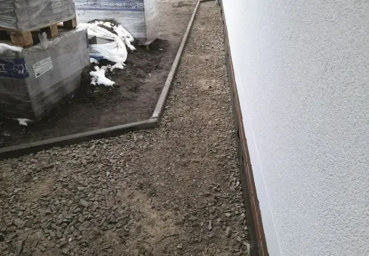 Ułożona opaska przeciwwysadzinowa z kruszywa wokół budynku, obok palety z materiałami budowlanymi.