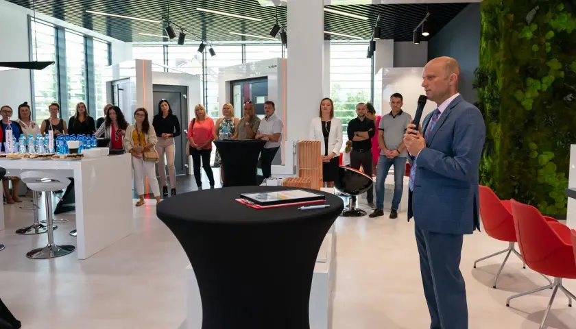Mężczyzna w garniturze przemawia do grupy osób w nowoczesnym salonie okien i drzwi.
