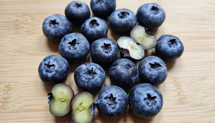 Kusząca garść borówek amerykańskich odmiany 'Bluecrop' i 'Duke' na drewnianej desce. Dwie jagody przekrojone ukazują soczysty, zielonkawy miąższ.