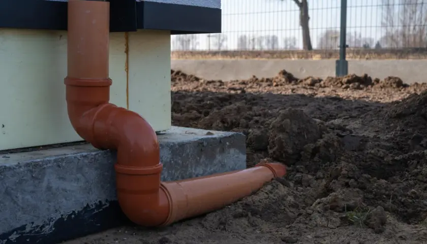 Pomarańczowe rury do odprowadzenia wody z rynien, połączone kolankami, biegną od ściany budynku w kierunku ziemi.